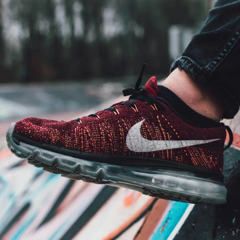 Nike athletic shoe with red Flyknit knit texture, silver Swoosh logo, and transparent Air Max sole, photographed against a blurred colorful background